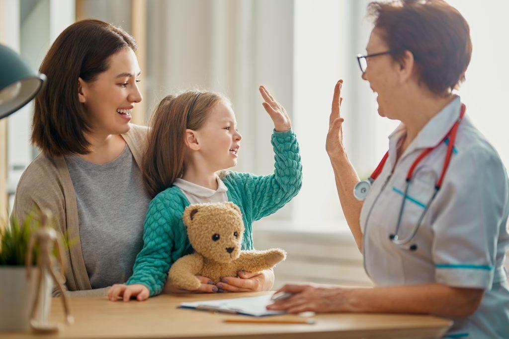 Mom holding a young girl in a green sweater who is high fiving a doctor as they sit at a table.