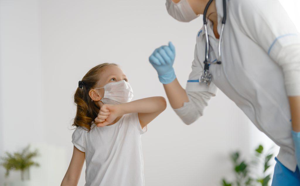 Doctor or nurse and a child bumping elbows with each other. Both of them are wearing masks.