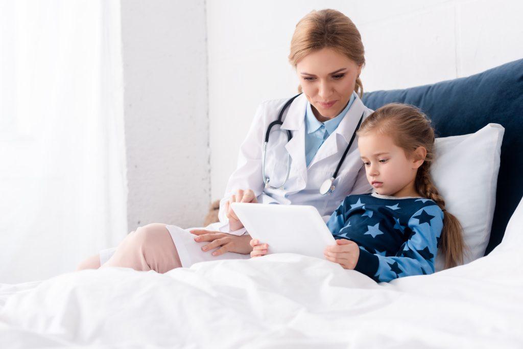 Doctor in white coat sitting near sick young girl with digital tablet